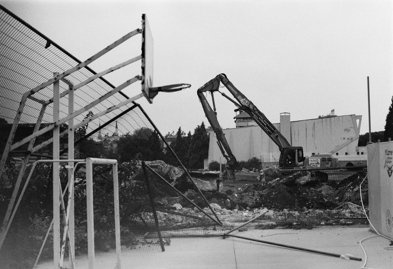 Black and white image of an urban demolition site featuring a dismantled basketball hoop and heavy machinery.